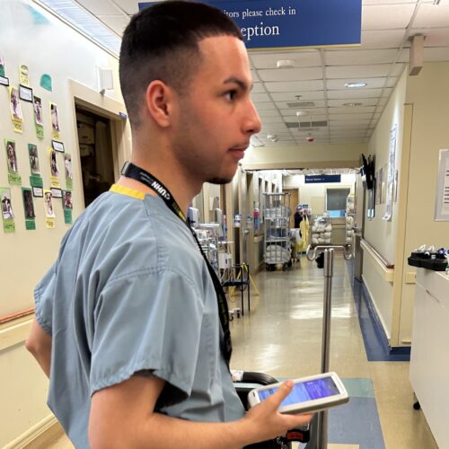 A young man wearing scrubs pushing a cart and using a handheld electronic device.