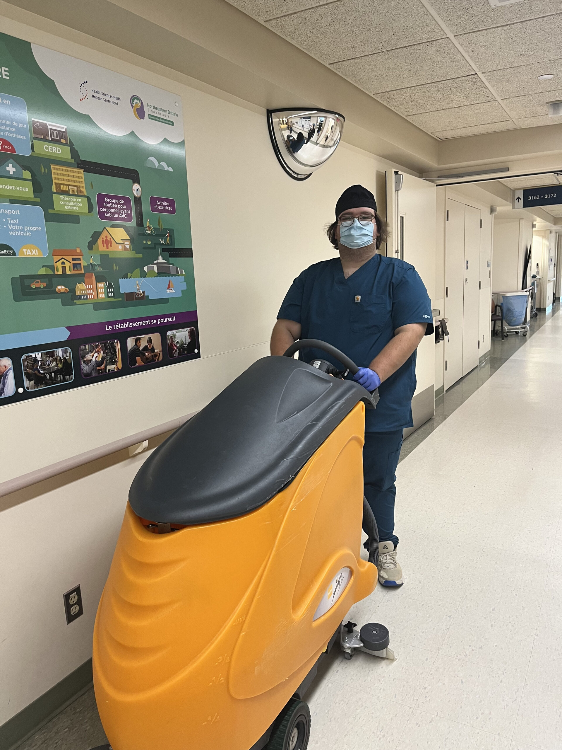 A man in a medical mask and scrubs pushing a yellow floor cleaning machine.