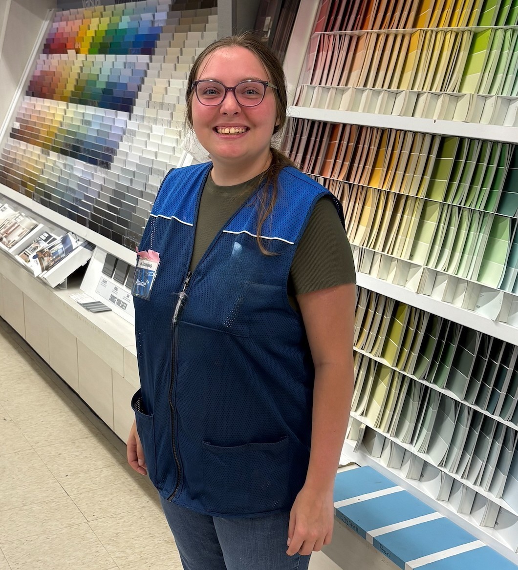 A woman wearing a blue vest standing in front of a wall of paint chips in a paint store.
