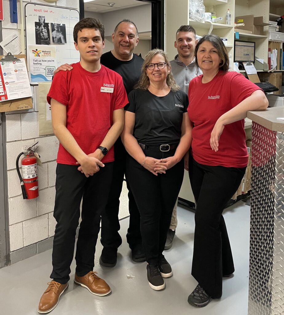 Five people in an office store room. A young man with a disability and an older woman are wearing red shirts, while the rest are wearing black shirts.